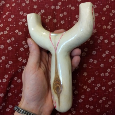 A smooth, varnished slingshot with pink linework and a shiny eye in the wooden handle, being held in front of a flower patterned fabric.