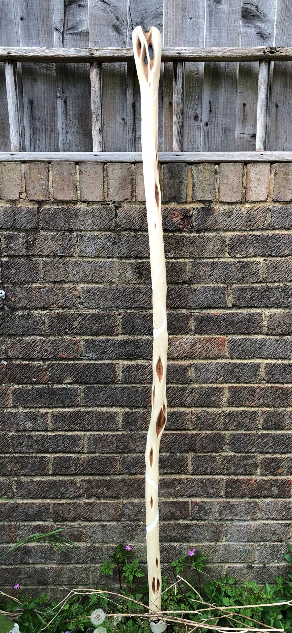 The full photo of the staff from the front, leaning up against a brick wall with wooden fencing above, and flowers in the garden below. It's white debarked wood with white curved lines wrapping around it, with diamond-shaped brown eyes on it, and a blue stone set into the branchy head at the top.