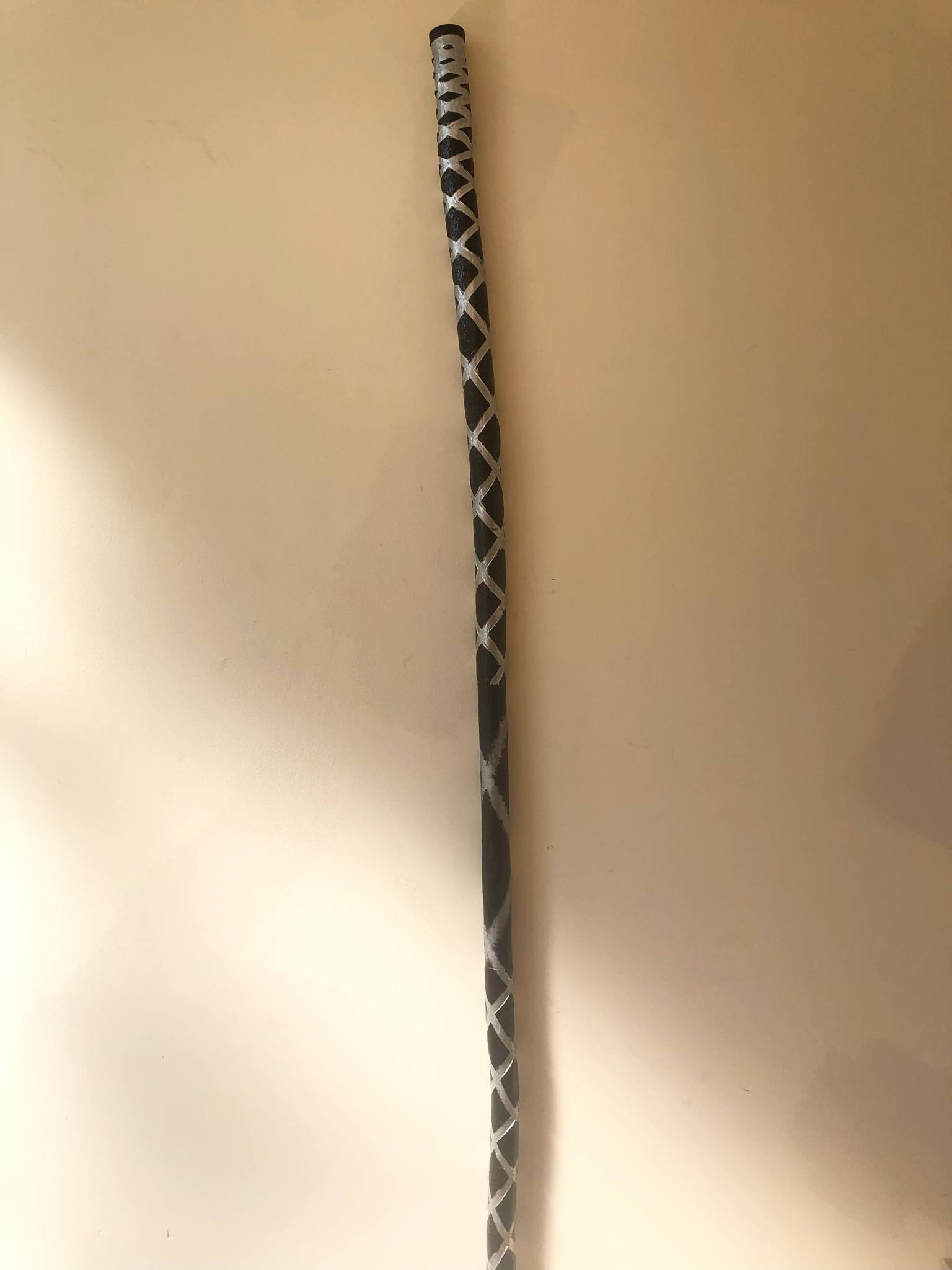 The Staff, painted black with an overpainting of the silver inside the grooves, leaning up against a wall, partially in the sun.