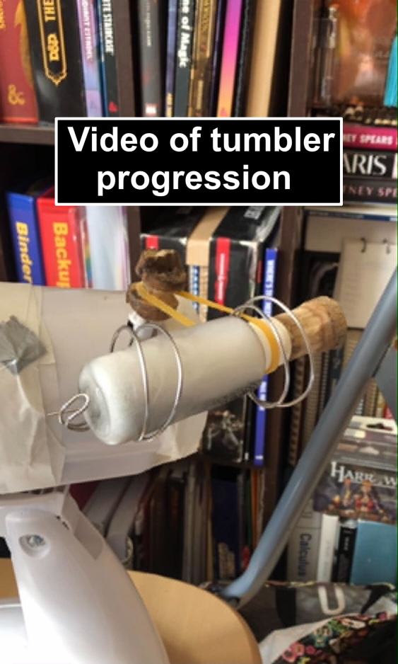 A pic of the final tumbler, with text saying video of tumbler progression. It begins as a simple cardstock tube cut into two pieces so that a rubber band can reach through the middle, taped to the side of a desk fan with a small rotating knob, and a small, clear plastic tube inside being rotated by a rubber band. It transitions to a photo of a similar setup, but with more tape and a less cut apart cardstock tube, and then another photo of the bottle being suspended by a bridge of wire looping around the bottle at the top and bottom, and the rubber bandgoing around a black electrical tape extension of the drive knob, followed by the running of the same wire setup, but with a larger wooden knob made from an old stick pinned above the drive knob.