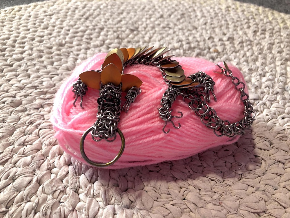 An orange and yellow striped dragon with hooks for claws, spreading one of his claws way out to show off the hooks, sitting on top of a spool of pink yarn, on top of a white crocheted rug.
