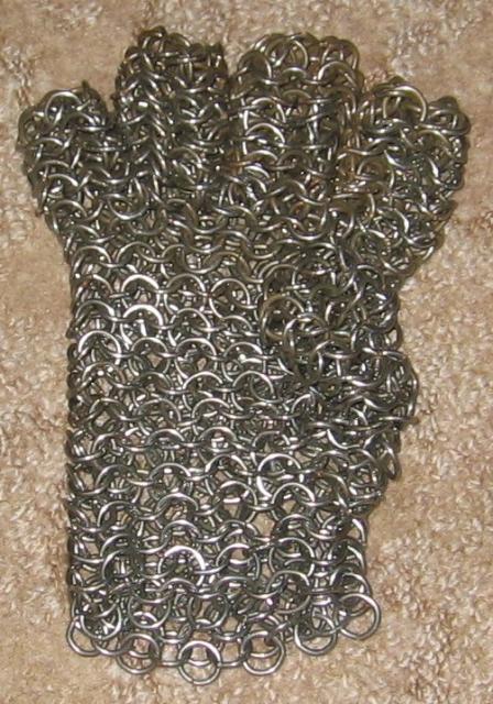 A rough looking heavy chainmaille glove laying on a light brown carpet. The rings are all very thick and dark, with some slight glinting off of some of them.