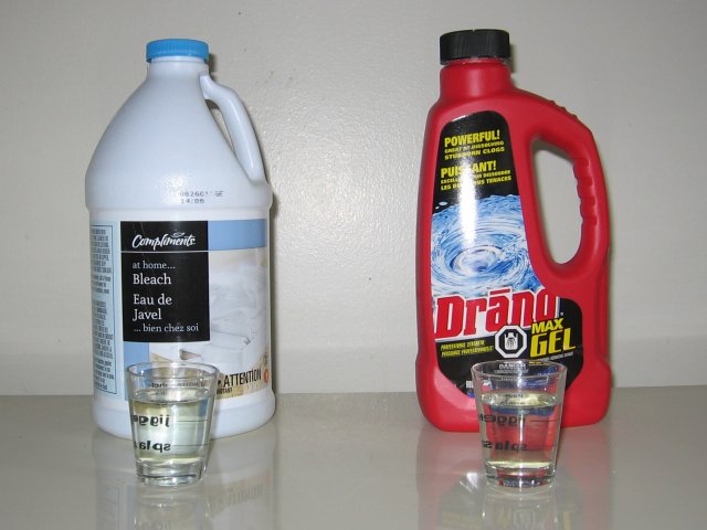 A bottle of bleach and draino with glasses in front on a kitchen counter.
