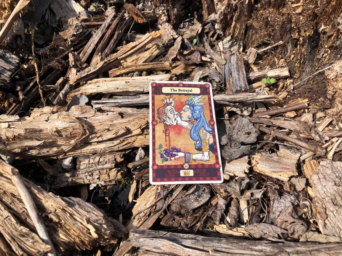 A woman kissing a severed head, the card laying on the forest debris.