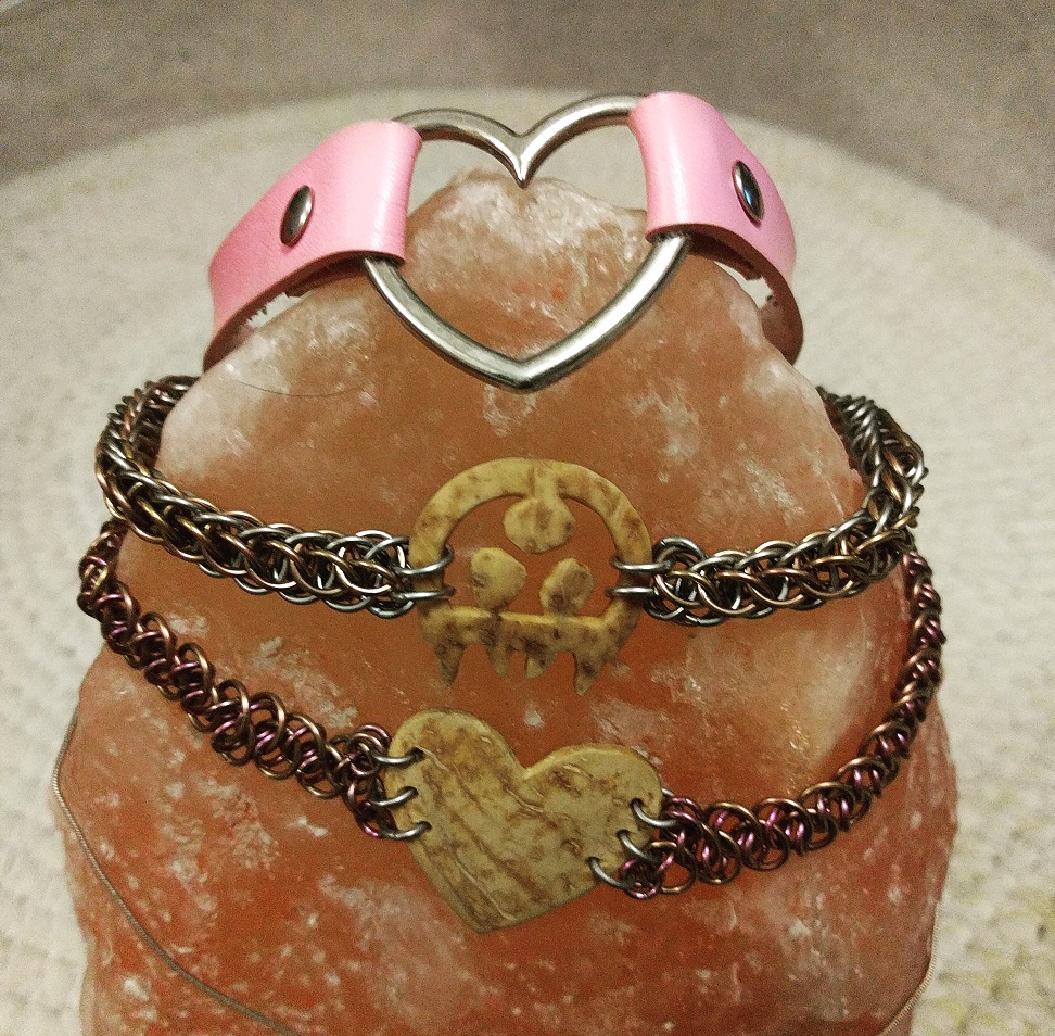 Three collars sitting on a himalayan salt lamp. One is a silver heart on a pink strap, one is a speckled brown Metroid with a somewhat round chainmaille strap, and one is a similar material heart with a pink and purple chainmaille strap.