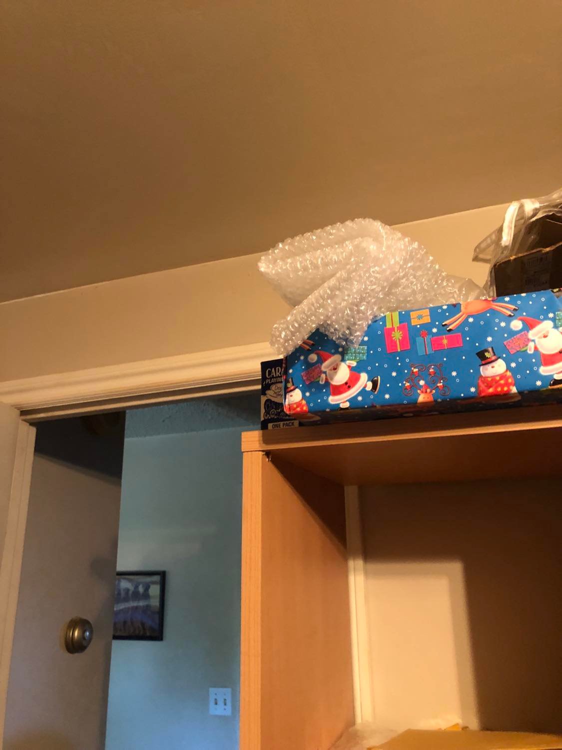 A blue card box, hidden beside a wrapped box and some bubblewrap on top of a shelving unit, though precariously placed, almost as though deliberately.