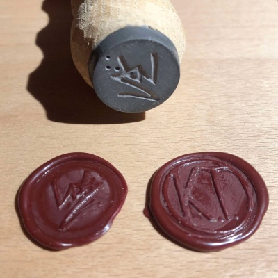 A pair of wax seals, one with a W Z logo and the other with a K dot T dot in a circle, in front of a dark grey W Z stamp on a wooden handle.