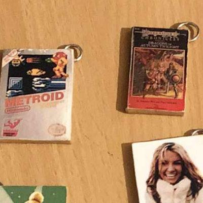 A closeup photo of several miniature ornaments made in the design of the original Metroid Nintendo box, the first edition release of Dragonlance Chronicles book 1, dragons of autumn twilight, and a Britney Spears ornament