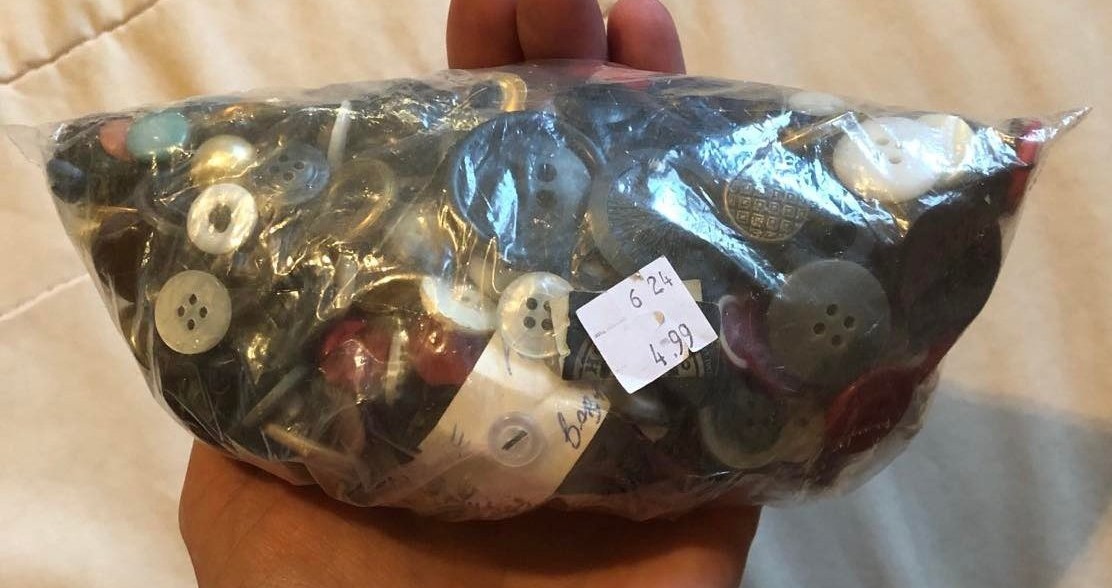 A pile of assorted buttons in a doubled clear plastic bag, with a 4.99 price tag on it. It's sitting in Kab's hand, with a white backdrop