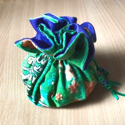 A green floral-patterned fabric pouch with a shimmering purple fabric interior fabric, closed and sitting on a wooden table.