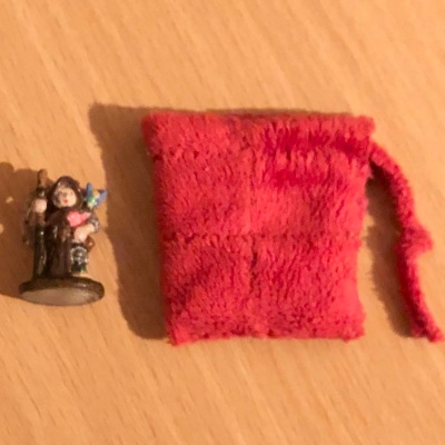 A tiny red pouch made from several patches of the same velvety material, beside a mini of a small humanoid holding a staff with a fairy on their shoulder, laying beside it.