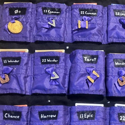A sheet of shimmery purple pockets on black fabric, each labelled different things like zero e, 13 common, tarot, harrow, 13 epic, and more, each with a drawstring tipped by a coin.