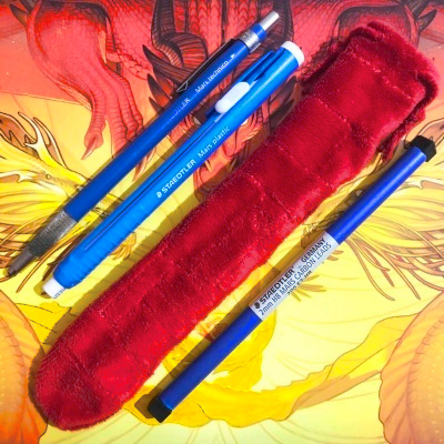 A red patchwork tube shaped bag, beside a pencil, stick eraser, and tube of spare pencil leads, on top of a red and gold dragon based backdrop.