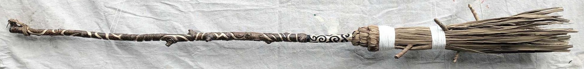 A homemade witch's broom with an intricately carved handle, curved linework running along the length of it, with bristles made from cabbage palm leaves. An extra swirly portion of the handle is just above the bristles, and a loop to store the broom is around the top.