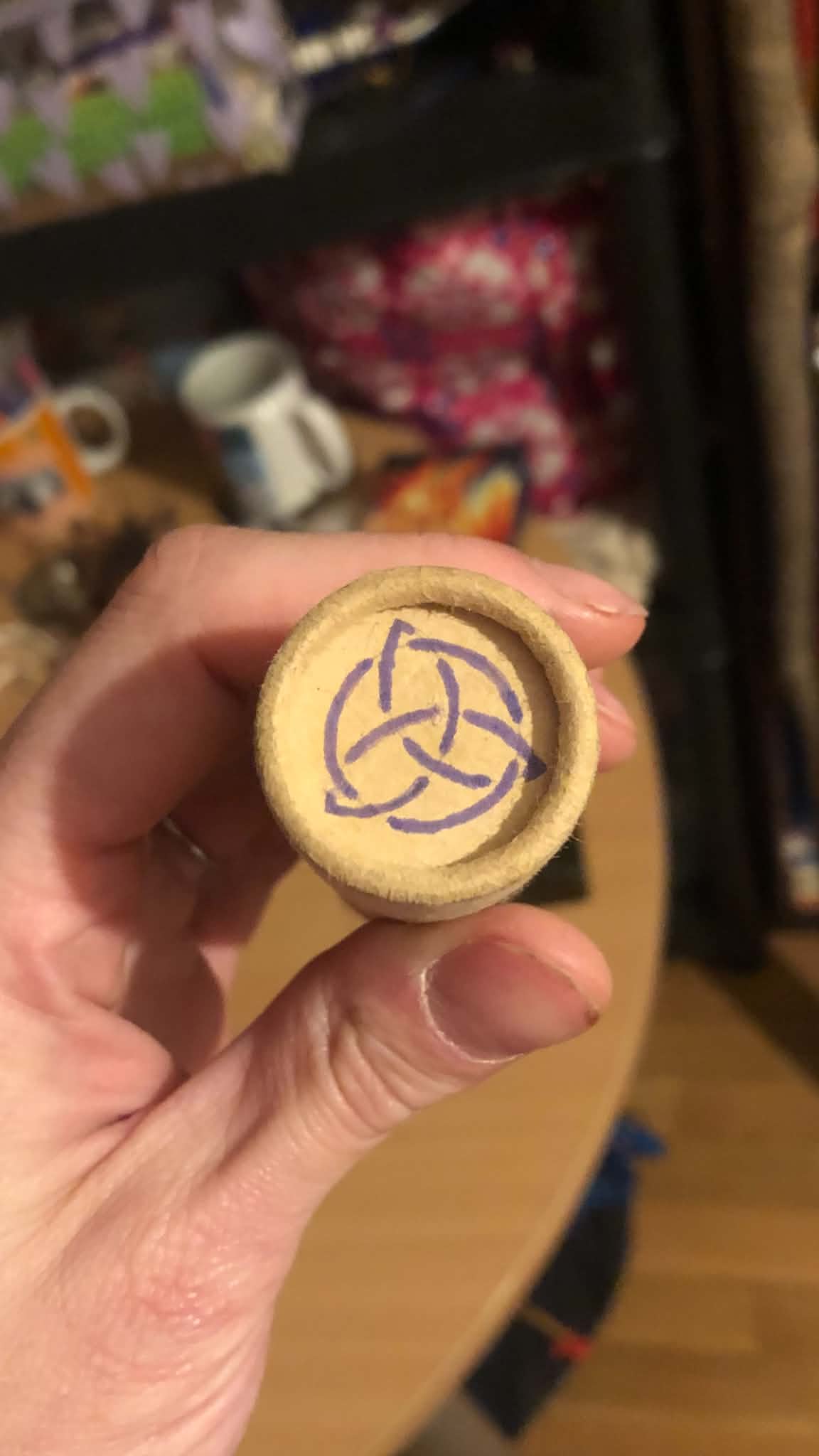 The lid of the thick paperboard tube that holds the casting sticks, with a light purple triquetera drawn onto it.
