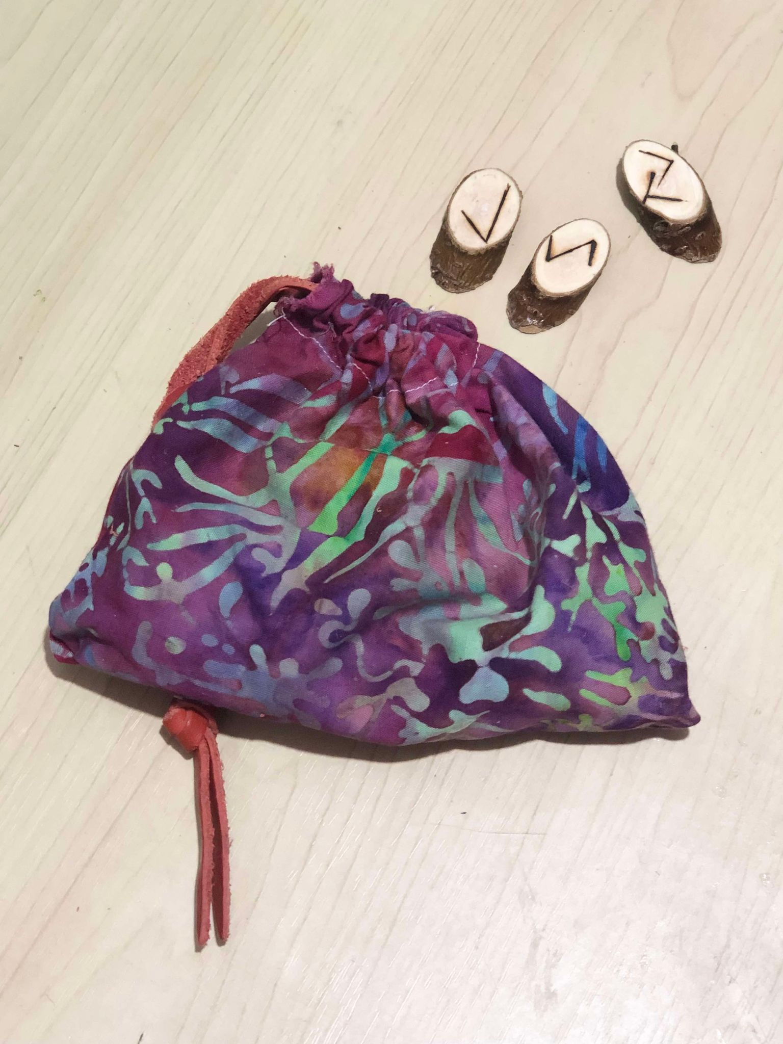 A multicoloured floral linework pouch, made up of purples, green, and blue, with an orange leather drawstring, with showing three runes from inside, thin disks of wood with the bark still around the sides, and runes put onto them with a woodburning kit.