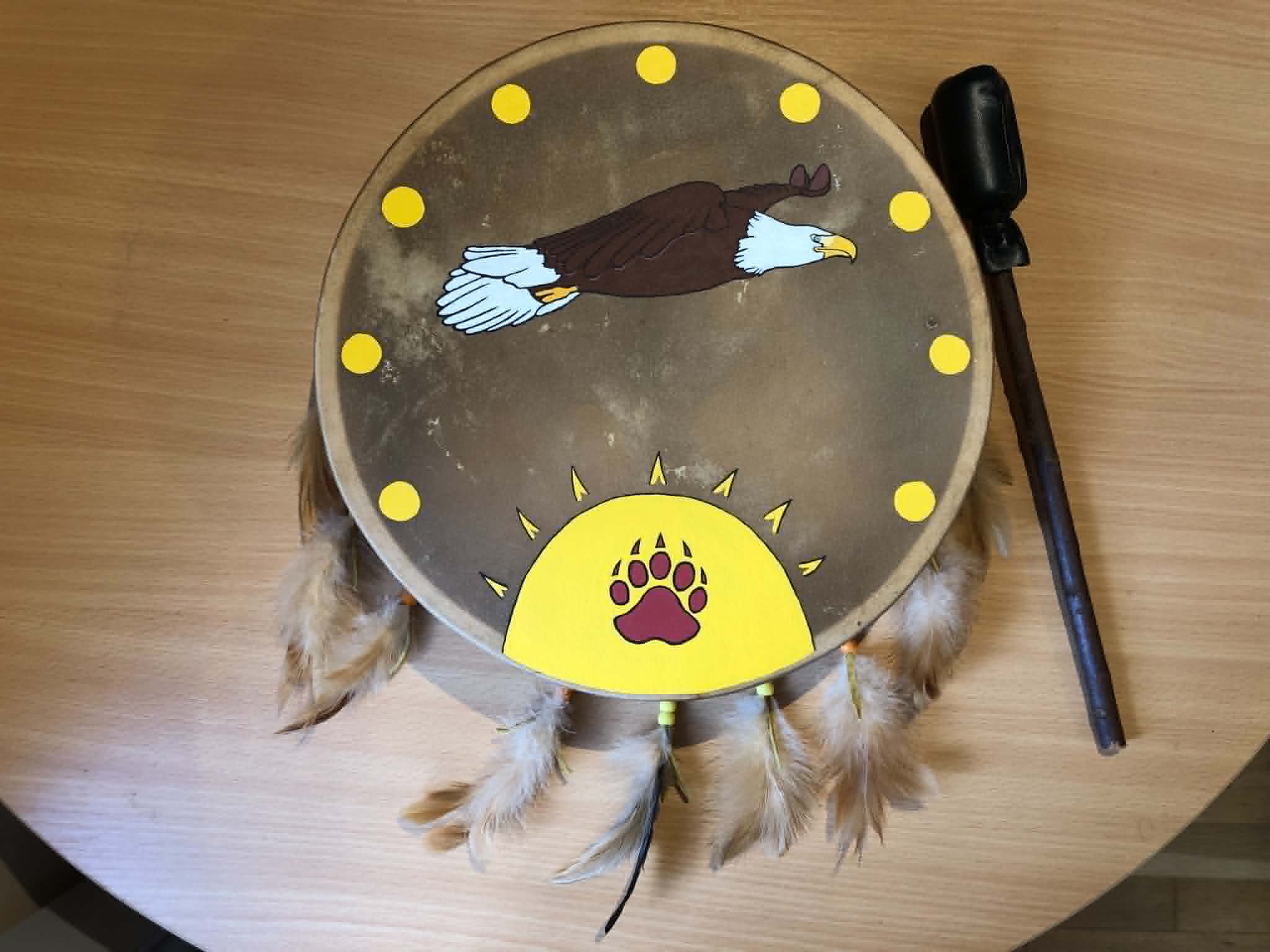 A leather hand drum with an eagle and suns motif on the face, and feathers attached all around.