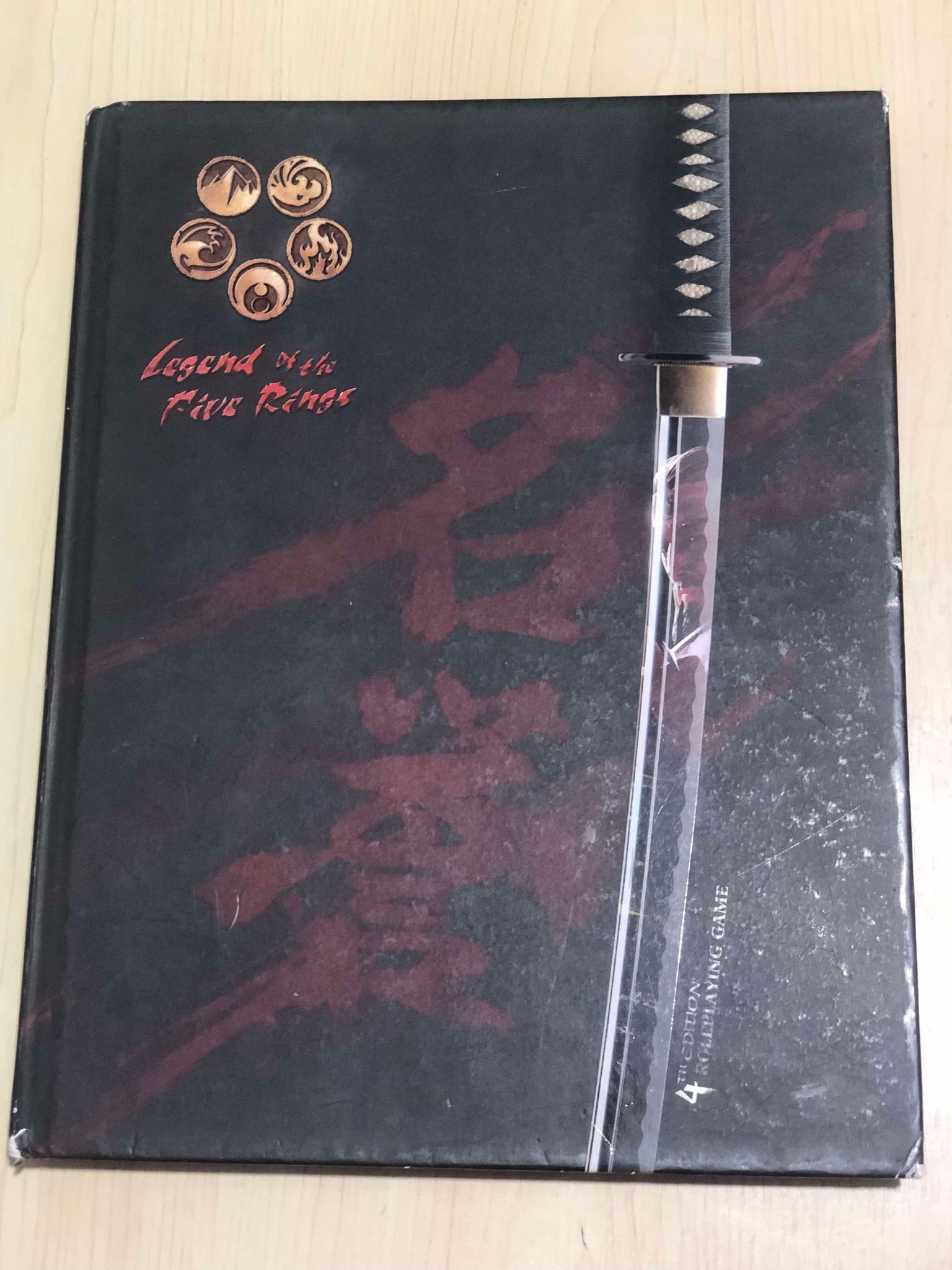 A hardcover book with primarily black with red accents, and a samurai sword prominently down the front, sitting on a wooden table, titled Legend of the Five Rings, fourth edition roleplaying game.