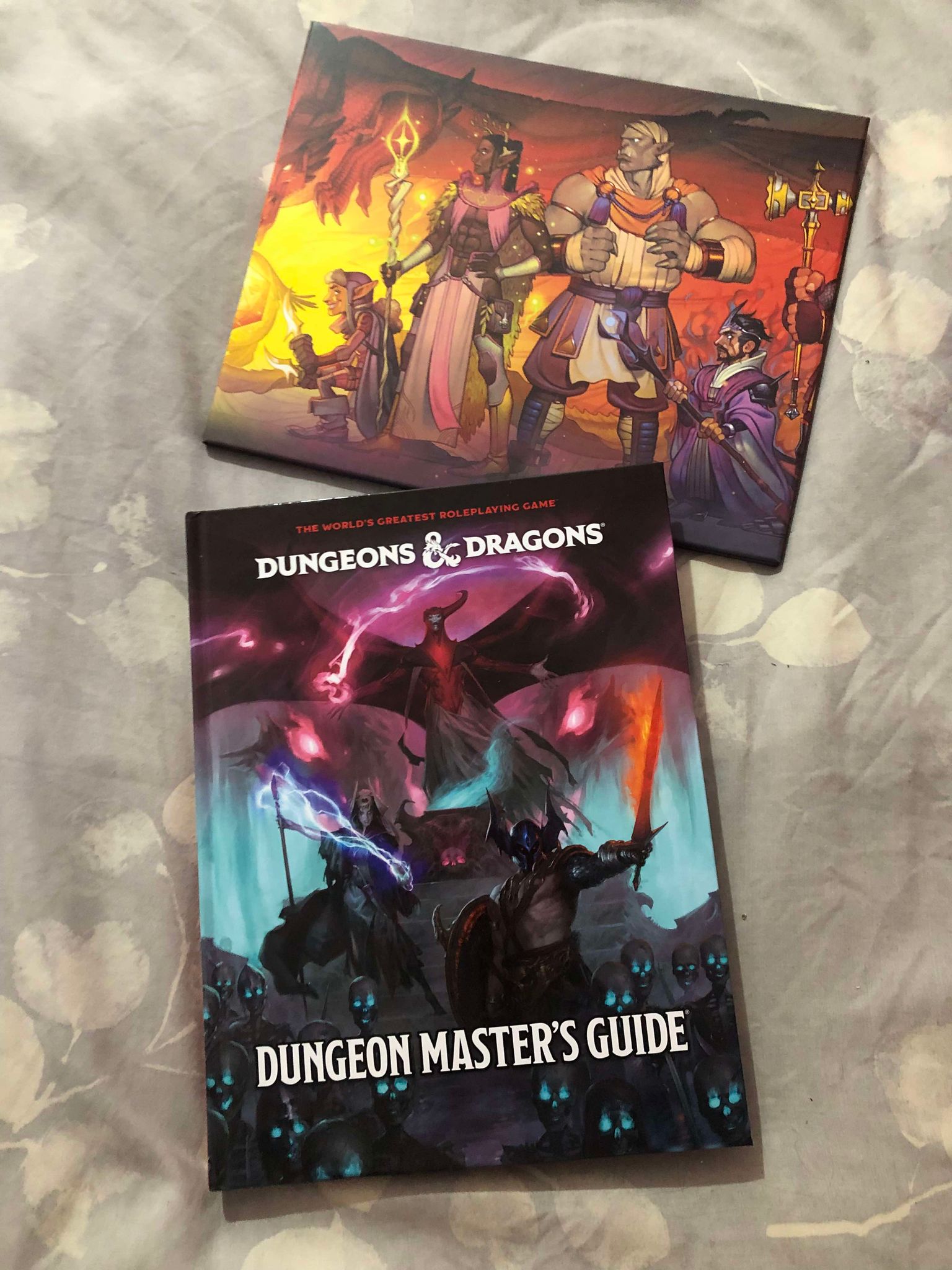 The book for the Dungeon Master's Guide and the 2024 Dungeon Master's Screen, the book having a cover of monsters, a horde of glowing, blue eyed skeletons, and the one horned winged monster Venger, while the dungeon master's screen shows a group of adventurers gathered around a friendly dragon ready to play.