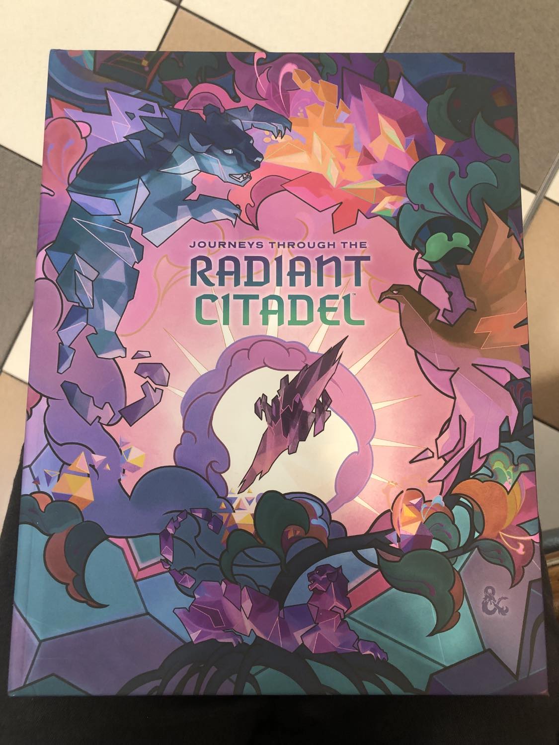 A book of Journeys through the Radiant Citadel, sitting on Kabutroid's lap in a mall with a checkered floor. It's the alternate cover, which shows somewhat polygonal style drawings of a jungle cat, feathered bird, clouds, and crystals, with in the center the radiant citadel itself, a huge crystal floating in space, enwrapped in a giant carved out fossil, all in pastel pinks, greens, and blues.