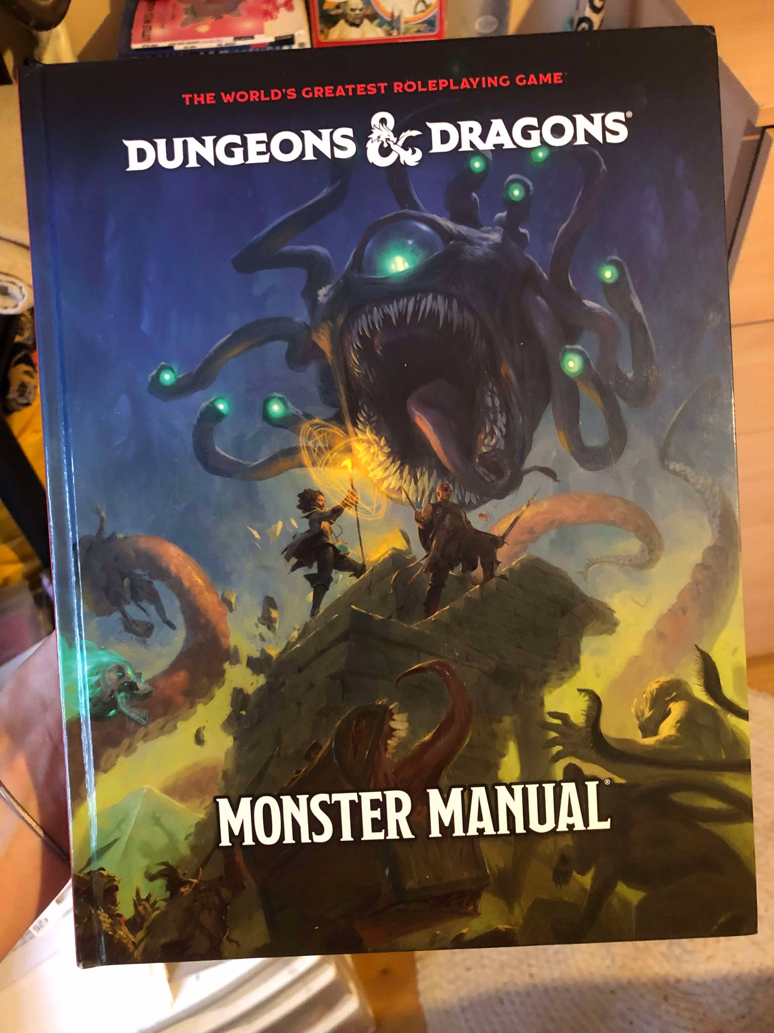Kabutroid holding the Monster Manual in front of her bookshelf. The cover is a murky blue backdrop, with a large, screaming beholder with glowing blue green eyes in front of a pair of heroes atop a crumbling tower, a third hero being pulled away by a giant orange tentacle which is further damaging the tower, and a glowing skull, chest mimic, and other monsters are all below, reaching up towards the remaining heroes from the ground.
