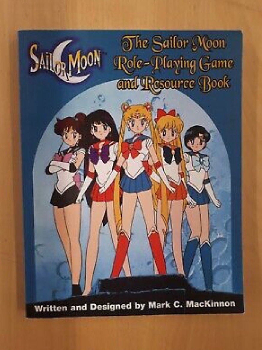 A blue book on a wooden table, titled The Sailor Moon Role-Playing Game and Resource Book, showing the five sailor scouts against a backdrop of the moon.