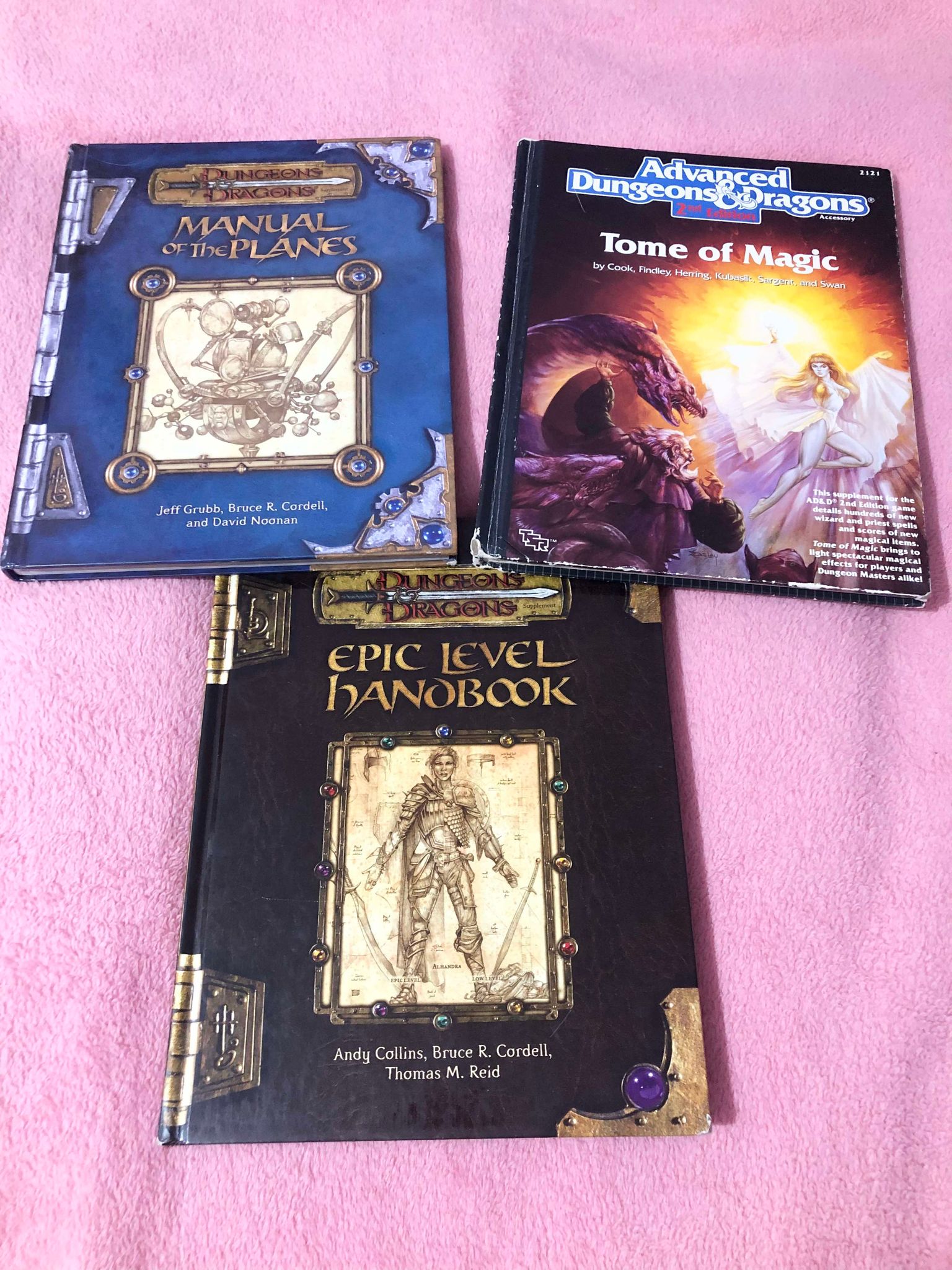 Three books on a pink blanket, fourth edition manual of the planes, second edition advanced dungeons and dragons tome of magic, and fourth edition epic level handbook.