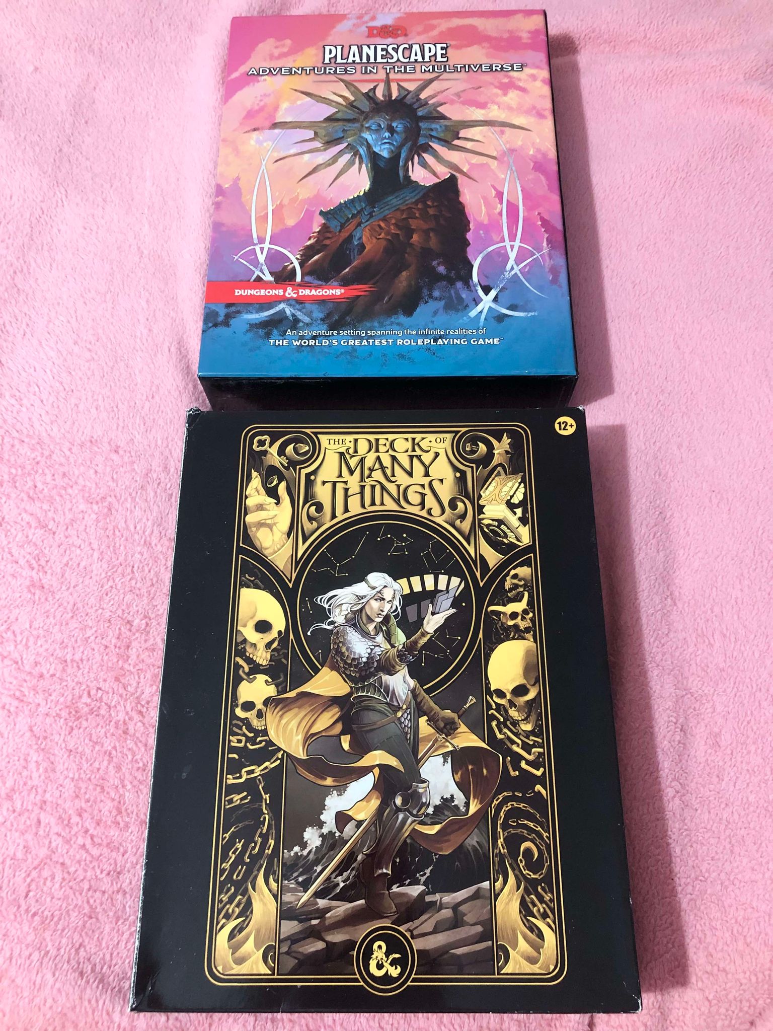 Two box sets sitting on a pink blanket, Planescape, adventures in the multiverse, and The Deck of Many Things, which came with a deck of 66 cards to go with it.