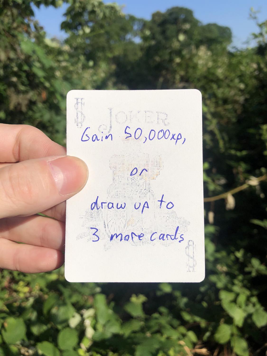 The coloured joker, held with the trees and foliage behind. Gain 50,000 xp, or draw up to 3 more cards.